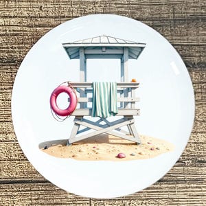 May include: A white ceramic plate featuring a beach scene. A lifeguard stand with a striped towel, a pink life preserver, and seashells on the sand. The stand is painted in light blue and white.