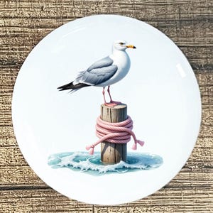 May include: A white ceramic plate featuring a detailed illustration of a seagull perched on a wooden piling wrapped with pink rope. The seagull has gray wings and a yellow beak, with ocean waves in the background.