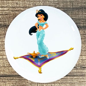 May include: A white round sticker with a cartoon image of Princess Jasmine from Disney's Aladdin. She is standing on a magic carpet with a golden lamp. She is wearing a blue and gold outfit with a tiara.