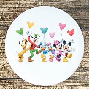 May include: White ceramic plate featuring a colorful illustration of Disney characters, including Mickey Mouse, Minnie Mouse, Donald Duck, Daisy Duck, Goofy, and Pluto. Each character is holding a balloon. The plate has a glossy finish.