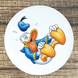 May include: White ceramic plate featuring a cartoon illustration of Donald Duck laughing hysterically. Donald Duck is depicted in his signature blue sailor suit, orange beak, and yellow feet. His blue hat is floating above him.