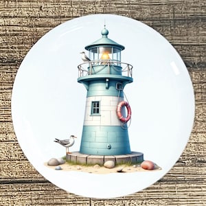 May include: A decorative plate featuring a lighthouse with a teal and white color scheme. A seagull perches on the railing, and another stands on the sand. A red life preserver hangs on the side of the lighthouse. Seashells are scattered around the base.