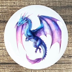 May include: A white, round decorative plate featuring a detailed illustration of a blue dragon with purple and pink wings. The dragon is in flight, with its wings spread. The artwork has a fantasy theme.