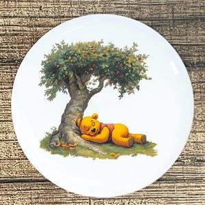 May include: A white, round plate featuring a sleeping yellow bear under a tree with red apples. The bear is wearing a red shirt and is lying on green grass. The tree has a brown trunk and green leaves.