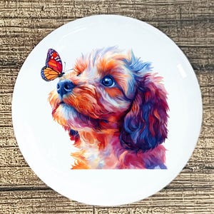 May include: A white ceramic plate featuring a colorful illustration of a puppy with a butterfly on its nose. The puppy's fur is rendered in shades of orange, red, blue, and purple. The butterfly has orange and red wings with black spots.