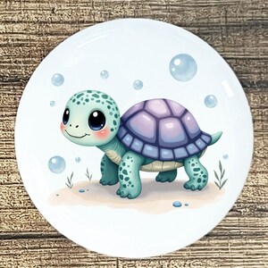 May include: A round, white ceramic plate featuring a cartoon turtle. The turtle has a light blue body with darker blue spots and a purple shell. Bubbles and small plants surround the turtle on a sandy surface.