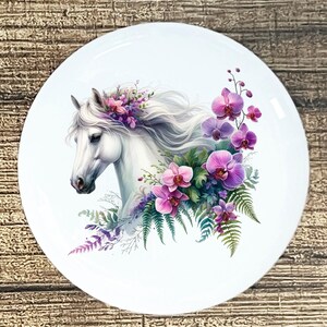 May include: White ceramic plate featuring a white horse head adorned with flowing white mane and a floral arrangement of pink and purple orchids and green ferns. The plate has a glossy finish.