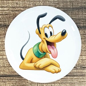 May include: A white ceramic plate featuring a cartoon illustration of Pluto, a yellow dog with black ears and a green collar. Pluto is smiling with his tongue sticking out. The plate has a glossy finish and is set against a wooden background.