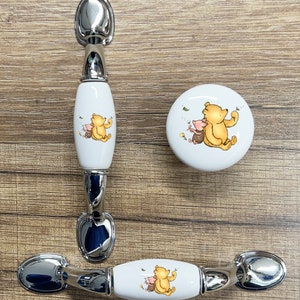 May include: Three ceramic cabinet knobs with a Winnie the Pooh and Piglet design. The knobs are white with a yellow and brown cartoon image of Winnie the Pooh and Piglet. Two of the knobs are rectangular and have a silver metal handle. The third knob is round and has no handle.