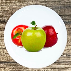 May include: Three apples on a white plate. One red apple, one green apple, and one red apple.