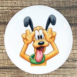 May include: A white ceramic plate featuring a cartoon illustration of Pluto, a yellow dog with black ears and a green collar. Pluto is making a funny face, with his tongue sticking out and fingers near his eyes.