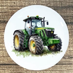 May include: A white ceramic plate featuring a watercolor illustration of a green tractor with yellow wheels. The tractor is depicted on a grassy field. The plate has a glossy finish and is set against a wooden background.