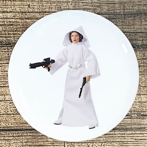 May include: A white plastic action figure of Princess Leia from Star Wars. She is wearing a white hooded robe and holding a blaster in one hand and a pistol in the other.