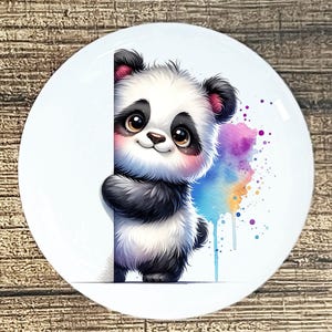 May include: A white ceramic plate featuring a cute panda bear illustration. The panda, with black and white fur, peeks from behind a white surface. Watercolor splashes of purple, blue, and pink add a colorful accent.