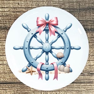 May include: A nautical-themed decorative plate featuring a light blue ship's wheel adorned with a pink bow and ribbons. The design includes seashells and a starfish on a sandy base, creating a coastal aesthetic.