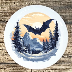 May include: White ceramic plate featuring a dark blue bat soaring over a snowy mountain landscape with pine trees. The background includes a sunset with orange and yellow hues. A decorative plate for Halloween or winter decor.