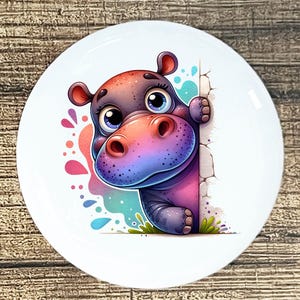 May include: A white ceramic plate featuring a cartoon hippo peeking from behind a wall. The hippo has a purple body, a pink and blue snout, and large blue eyes. Colorful paint splatters and green grass are also visible.