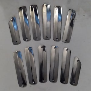 May include: A collection of twelve polished, rectangular metal pieces with a rounded top and a small hole near the top. The pieces have a shiny, reflective surface and are arranged on a light-colored background.