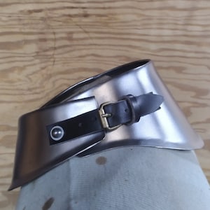 以下が含まれることがあります： 銀色の金属製の首ガード。黒い革のストラップと真鍮製のバックルが付いています。ストラップには、白い中心を持つ装飾的なリベットが付いています。