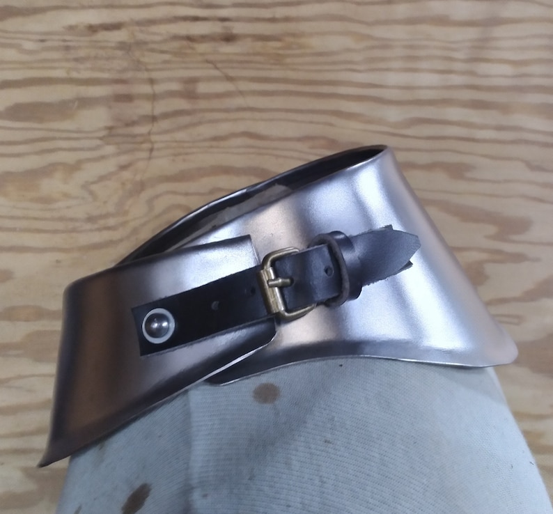 以下が含まれることがあります： 銀色の金属製の首ガード。黒い革のストラップと真鍮製のバックルが付いています。ストラップには、白い中心を持つ装飾的なリベットが付いています。