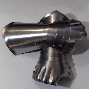Könnte beinhalten: Ein polierter Stahlhandschuh, wahrscheinlich für historische Nachstellungen oder Ausstellungen. Der Handschuh hat bewegliche Platten und Nieten, mit einer abgerundeten Hand und einer ausgestellten Manschette. Das Metall hat eine reflektierende Oberfläche, was auf eine gute Pflege hindeutet.