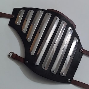May include: A black leather arm guard featuring seven vertical, polished silver-tone metal plates secured with rivets. The protective piece includes brown leather straps with metal buckles for fastening, providing a structured design for the forearm.