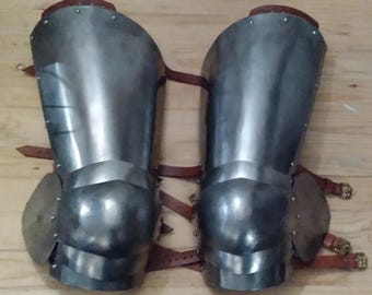 May include: A pair of steel knee armour with brown leather straps. The armour is made of metal plates and has a rounded shape. The straps are adjustable and have buckles.