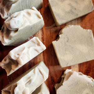 Puede incluir: Vista cenital de varias barras de jabón hechas a mano con un aspecto texturizado y orgánico. Los jabones son de varios tonos de verde pálido y blanco, con remolinos e imperfecciones visibles. Descansan sobre una superficie de madera oscura.