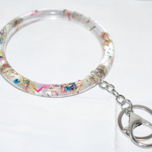May include: A clear, round keychain with colorful flower petals embedded inside. The keychain has a silver metal ring and a silver chain attached.