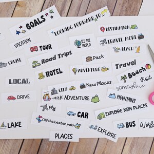 May include: A selection of travel-themed stickers featuring illustrations and text such as "Goals," "Vacation," and "Explore." A pair of pink scissors sits on the white paper, indicating a DIY project. The stickers are on a wooden surface.