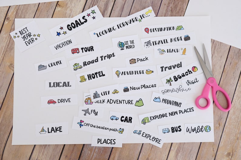 May include: A selection of travel-themed stickers featuring illustrations and text such as "Goals," "Vacation," and "Explore." A pair of pink scissors sits on the white paper, indicating a DIY project. The stickers are on a wooden surface.
