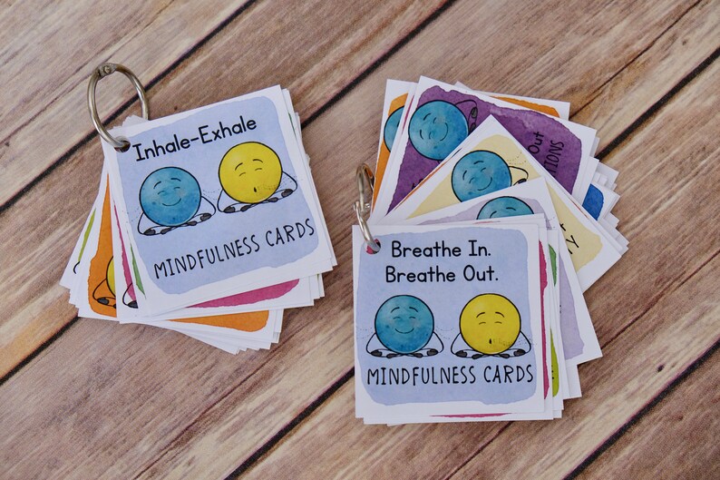 Puede incluir: Un juego de tarjetas de atenci&oacute;n plena de colores con ilustraciones de caras sonrientes y el texto "Inhala-Exhala" y "Inhala. Exhala." Las tarjetas est&aacute;n unidas con un anillo plateado.