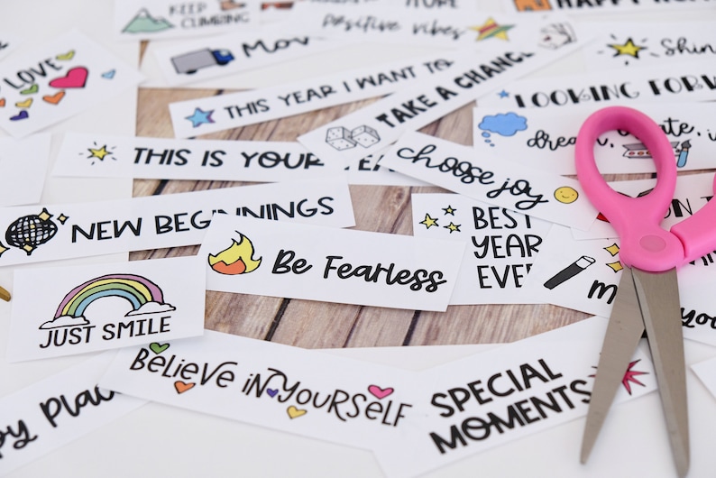 May include: A selection of white paper strips featuring handwritten motivational quotes and colourful illustrations, including rainbows and hearts. A pair of pink scissors lies on the papers, indicating a craft or DIY project.