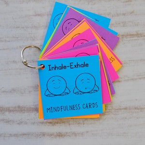 Puede incluir: Un juego de tarjetas de mindfulness coloridas con un archivador de anillas. La tarjeta frontal es azul y presenta una ilustraci&oacute;n de dibujos animados de una persona que inhala y exhala con el texto "Inhale-Exhale" y "Mindfulness Cards".