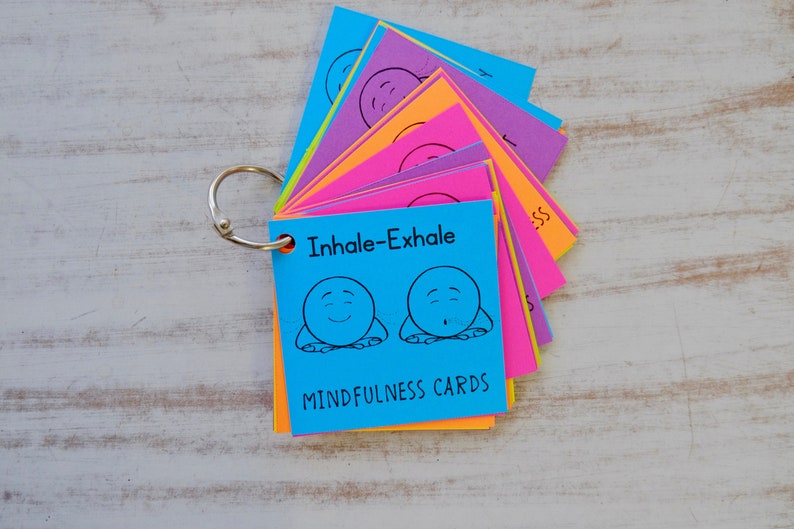 Puede incluir: Un juego de tarjetas de mindfulness coloridas con un archivador de anillas. La tarjeta frontal es azul y presenta una ilustraci&oacute;n de dibujos animados de una persona que inhala y exhala con el texto "Inhale-Exhale" y "Mindfulness Cards".