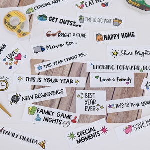 May include: A selection of white rectangular stickers featuring phrases and illustrations such as "Crafts," "Get Outside," "Happy Home," and "Save Money." Gold scissors and a roll of Scotch tape are also visible.