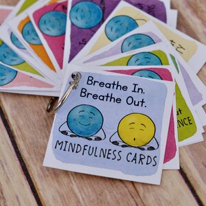 Puede incluir: Un juego de tarjetas de mindfulness coloridas con un personaje de dibujos animados azul y amarillo en la tarjeta frontal. Las tarjetas est&aacute;n unidas con un anillo plateado. El texto en la tarjeta frontal dice "Breathe In. Breathe Out. Mindfulness Cards."