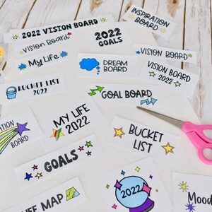 May include: A selection of white cards with handwritten text and illustrations, such as "Vision Board 2022" and "Dream Board." A pair of pink scissors and a roll of tape are also visible, alongside the cards.