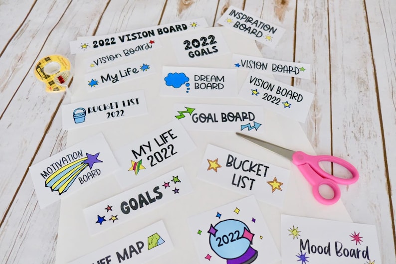 May include: A selection of white cards with handwritten text and illustrations, such as "Vision Board 2022" and "Dream Board." A pair of pink scissors and a roll of tape are also visible, alongside the cards.