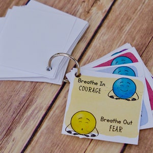 Puede incluir: Un conjunto de tarjetas coloridas con caras de dibujos animados y el texto "Breathe In COURAGE" y "Breathe Out FEAR". Las tarjetas est&aacute;n unidas por un anillo de metal.