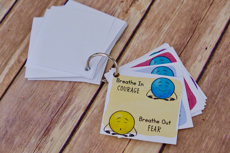 Puede incluir: Un conjunto de tarjetas coloridas con caras de dibujos animados y el texto "Breathe In COURAGE" y "Breathe Out FEAR". Las tarjetas est&aacute;n unidas por un anillo de metal.