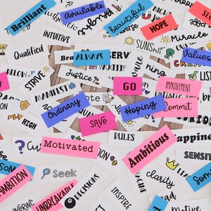 May include: A selection of white rectangular stickers with words and phrases such as "Ambition", "Hope", and "Dreams" in black ink. Some stickers are pink and blue. Scissors and tape are also visible.