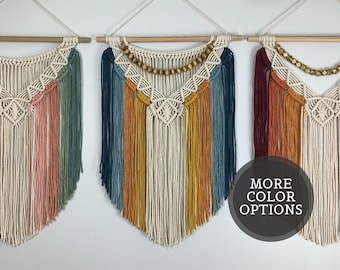 Pode incluir: Três tapeçarias de macramé penduradas em varões de madeira. Cada peça apresenta padrões de nós intrincados, franjas longas e contas de madeira. Os designs incorporam várias paletas de cores, incluindo rosa, verde, azul, amarelo e vermelho escuro.