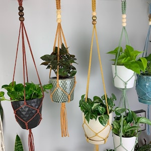 May include: Five hanging plant macrame planters with different colored cords and plants. The planters are made of natural fibers and have a bohemian style. The plants are in various pots and include a green plant, a leafy plant, a vine, and a succulent.