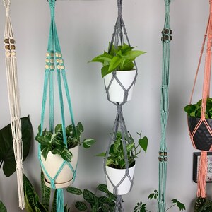 May include: Five macrame plant hangers in cream, turquoise, gray, mint green, and coral. The hangers feature knotted cord designs, some with wooden beads, holding various potted plants in white and black containers against a plain wall.