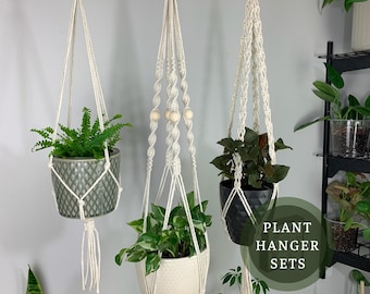Pode incluir: Três suportes de plantas em macramé branco-sujo suspensos do teto, cada um com uma planta em vaso. Os suportes apresentam designs de corda com nós e contas de madeira. O texto na imagem diz: PLANT HANGER SETS. Os vasos são verdes, brancos e pretos.