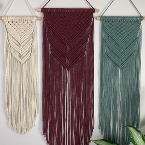 May include: Three macrame wall hangings suspended from wooden dowels. Each piece features a chevron pattern created with intricate knots and long, loose fringe. The hangings are colored in cream, deep burgundy, and muted sage green respectively.