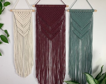 May include: Three macrame wall hangings suspended from wooden dowels. The left piece is cream, the center is deep burgundy, and the right is sage green. Each features a chevron-patterned knot design with long, loose fringe hanging below.