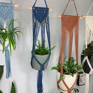 May include: Four macrame plant hangers suspended from wooden dowels, featuring intricate knotted cord designs in light blue, dark blue, terracotta, and cream. Each hanger supports a potted plant, with the blue and cream versions featuring wooden beads.