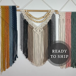 May include: Three macrame wall hangings in a row. The first is a gradient of cream, yellow, orange, blue, and navy. The second is a cream, gray, and brown design with wooden beads. The third is cream, pink, green, and navy. All are suspended from wooden dowels.
